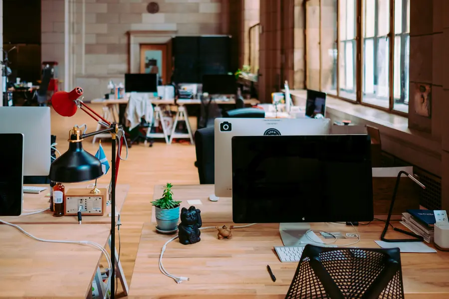 Desks with monitors and lamps along a row of tall windows